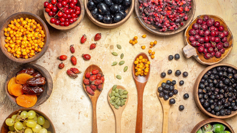 Photo comprenant plusieurs récipients et cuillères en bois, remplis de différents super-aliments comme des dattes, des graines, des raisins et des baies.